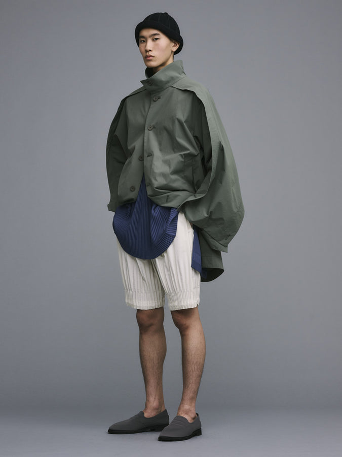 Model wearing WING COAT in green with blue top and off white shorts. Grey loafers and a black hat. Posing with their hands in their pockets.