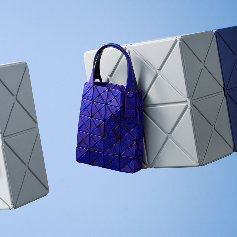PRISM PLUS small blue tote bag floating in the air next to grey cubes with the triangular pattern of BAO BAO ISSEY MIYAKE. Light blue background.