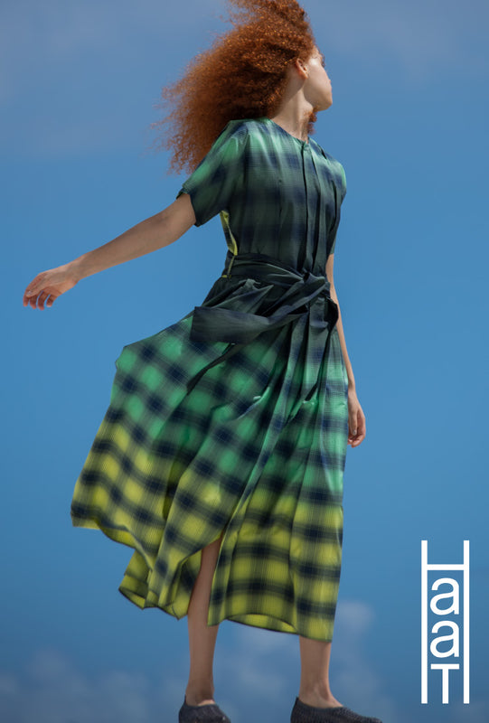 Model wearing GRADATION CHECK dress in yellow and green and posing against a backdrop of blue sky. Head facing backwards and right arm stretched out behind them. Bottom Right Corner: HaaT logo in white.