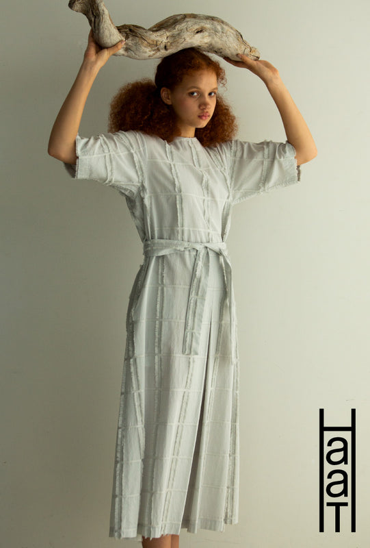 Model wearing YUUKI COTTON short-sleeved dress in grey. Shot from hips up, holding a log above their head. Bottom right: HaaT logo in black on white background.