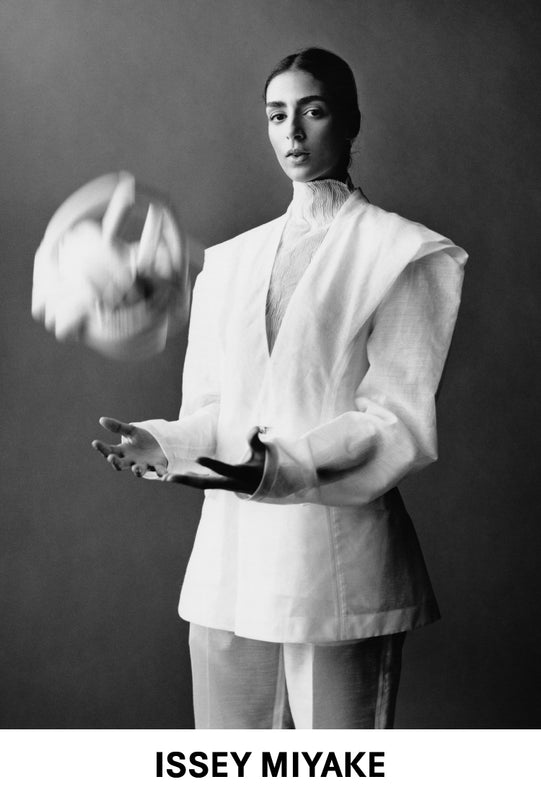 Model wearing TRANSLUCENT SUIT jacket and trousers in white. Photo is black and white and shot from their thighs up. Model is throwing a BREAD ACC bag in white in the air. Bottom: ISSEY MIYAKE logo in black on white background.