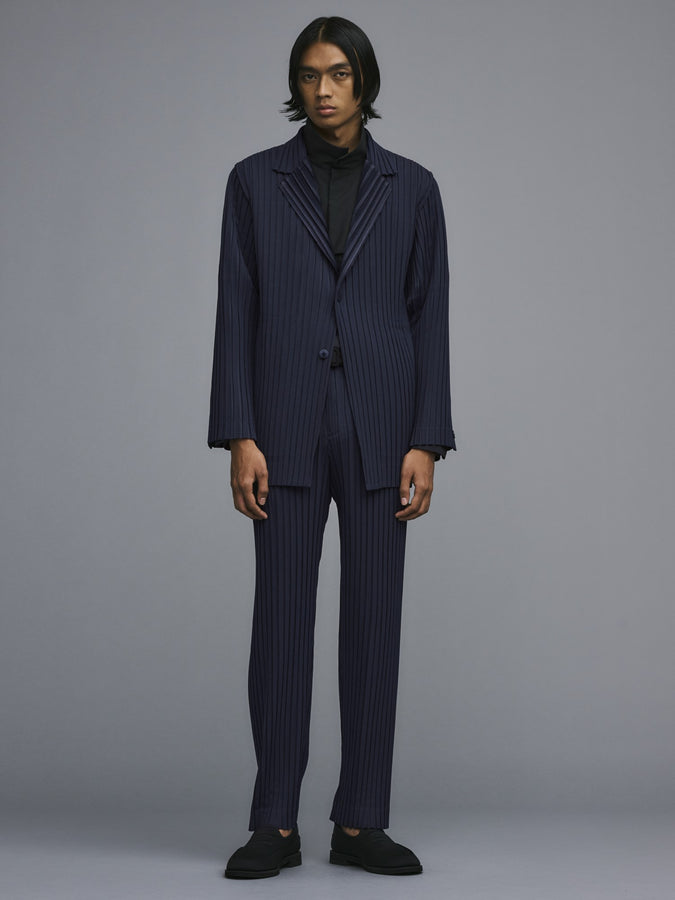 Model wearing BOX PLEATS ENSEMBLE suit in navy, paired with black loafers. Jacket worn over a black shirt with a stand collar. Posing against a grey background.
