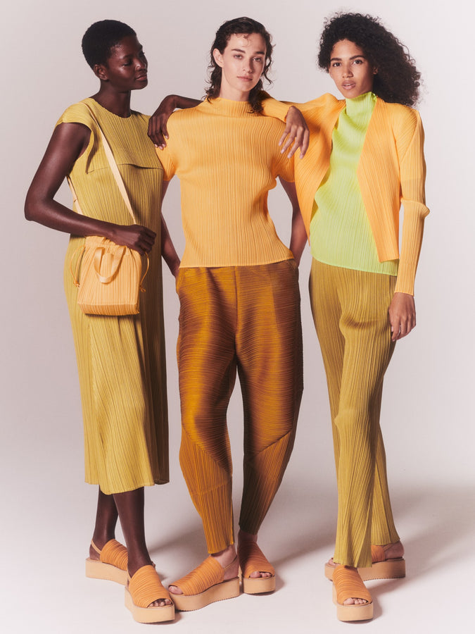 Three models wearing PLEATS PLEASE ISSEY MIYAKE clothing in orange, lime and yellow tones with their arms around each other.