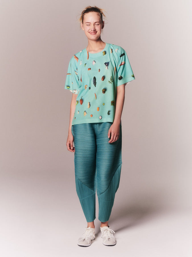 Smiling model wearing turquoise green blue clothes, MENU-T printed T-shirt and THICKER BOUNCE tapered trousers with white sneakers, posing against a beige background.