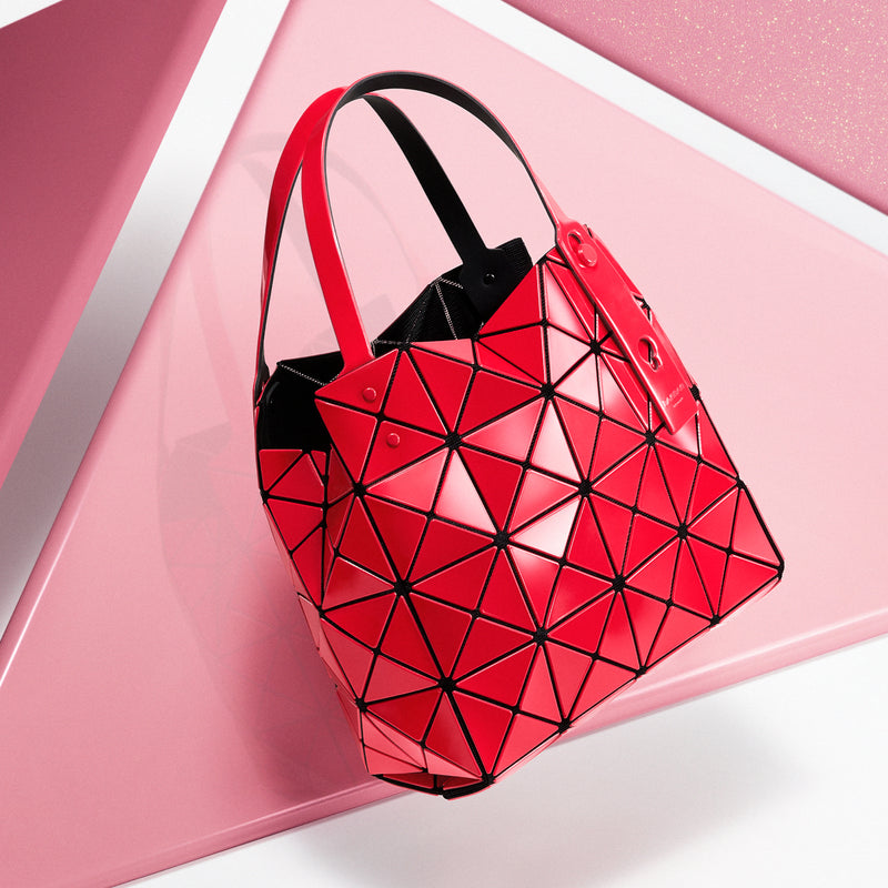 Red CARAT handbag floating mid air against backdrop of large pink triangles and a white background.