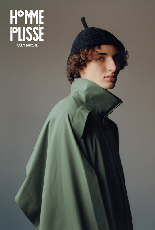 Top left corner: HOMME PLISSÉ ISSEY MIYAKE logo in white. Main: Closeup of model shot from elbows up, wearing the green WING COAT and black hat standing with body facing to the side but head facing to the front.