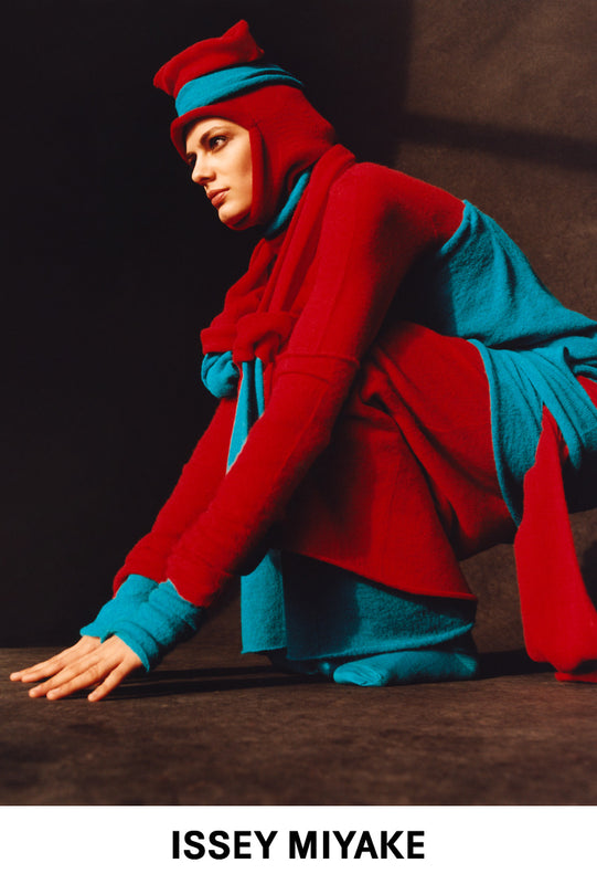 Top: Model crouched down wearing blue and red INTERPLAY dress under matching tunic and beanie with a red balaclava underneath. Bottom: ISSEY MIYAKE logo in black on white background. 