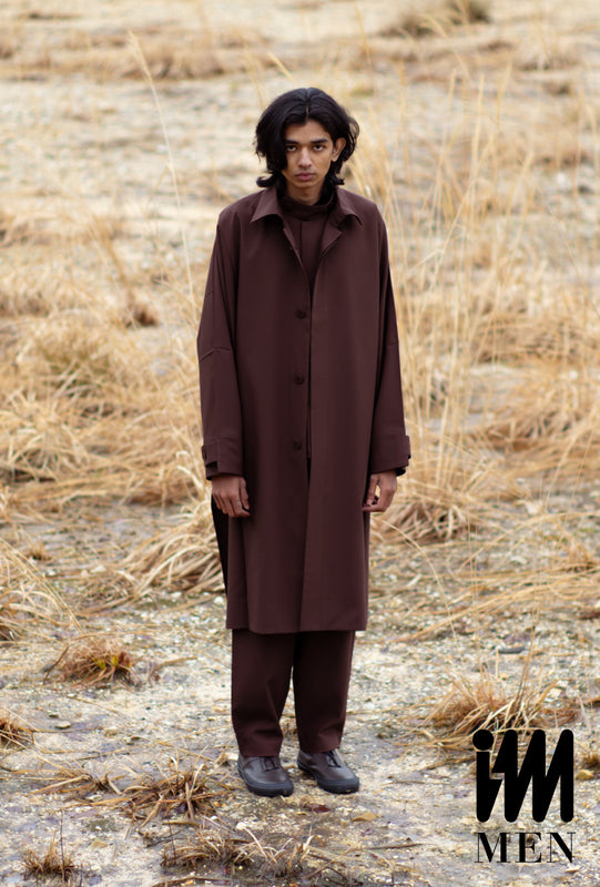 Main: Model wearing brown jacket, trousers, shirt and shoes standing in a field of dry grass. Slightly looking up at camera. Bottom right corner: IM MEN logo in black.