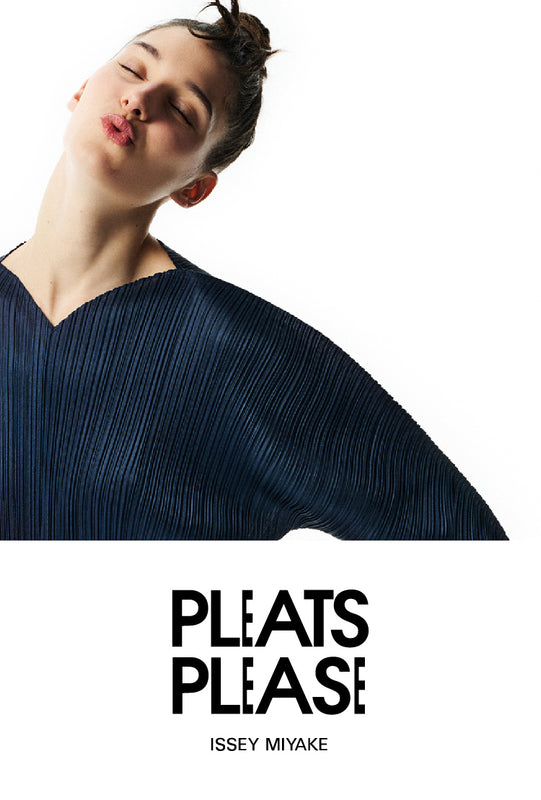 Top: Model wearing navy blue top with curved V-neckline and long sleeves, posing with hands on hips, head tilted to the left and puckered lips. Off white background. Bottom: PLEATS PLEASE ISSEY MIYAKE logo in black on white background.