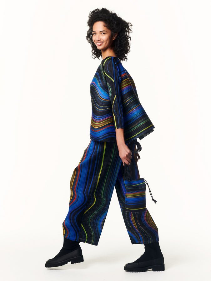 Model wearing black, blue, green and orange striped WARP top and matching trousers with black boots. Holding a matching striped bag. Shot whilst walking against a white background.