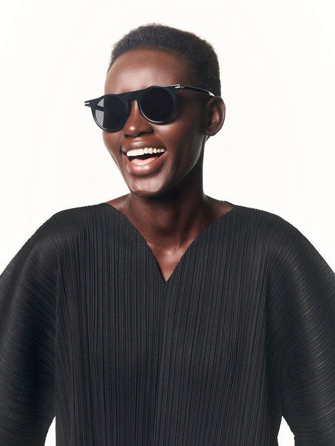 Model wearing black dress with V-neckline paired with black sunglasses. Shot from waist up. Model is smiling. White background.